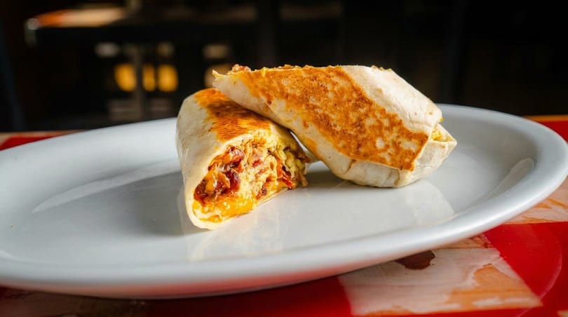 Two golden-brown breakfast burritos, one cut open to reveal eggs, cheese, and bacon, on a white plate.