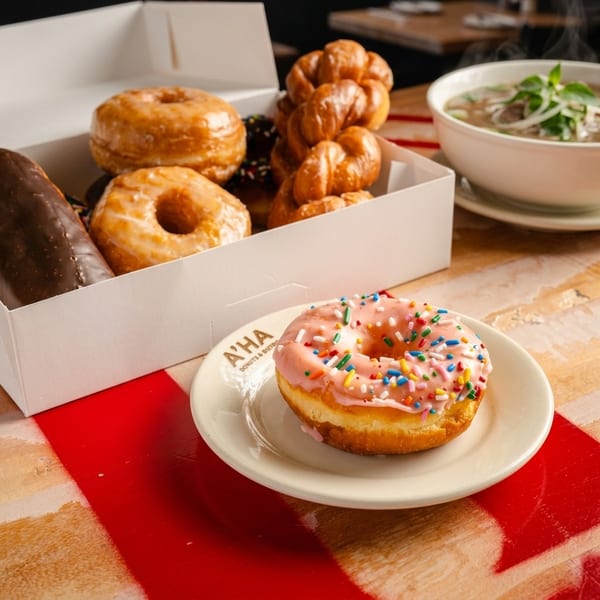Pink-frosted donut with sprinkles on a white plate, with a box of assorted donuts behind it.
